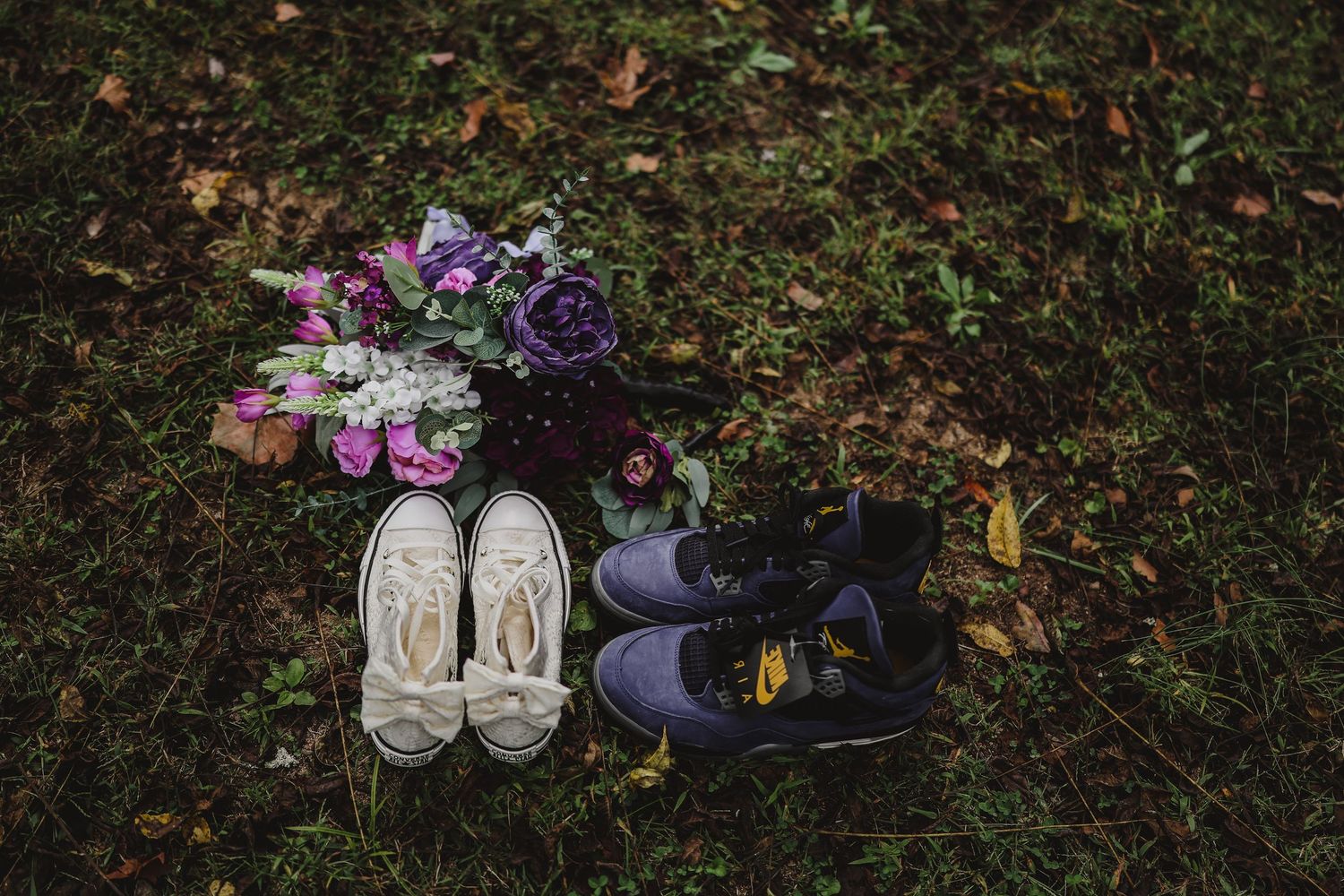 Cream sneakers and dark purple sneakers beside a bouquet of pink, white and purple flowers on grass with fallen autumn leaves.