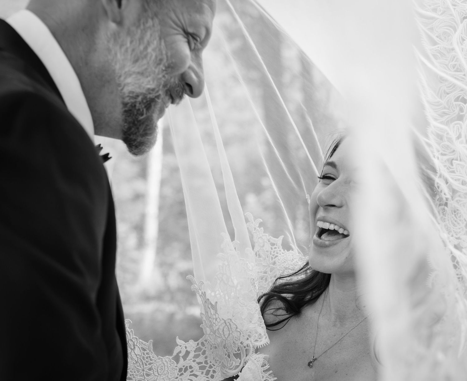 Black and white wedding photo of smiling bride in lace veil and groom sharing a joyful intimate moment.