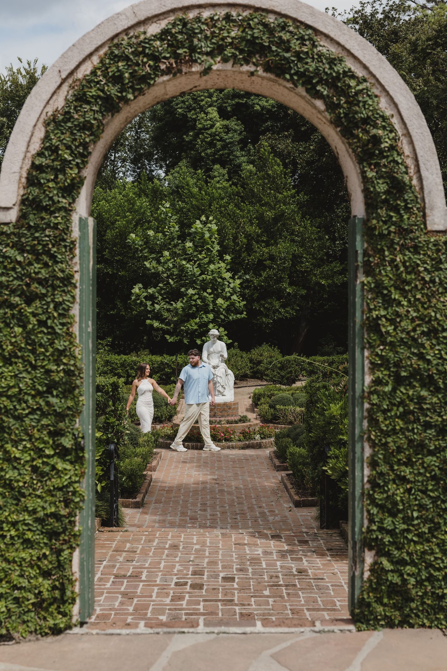 Couple walking hand-in-hand through a lush green garden archway on a brick path.