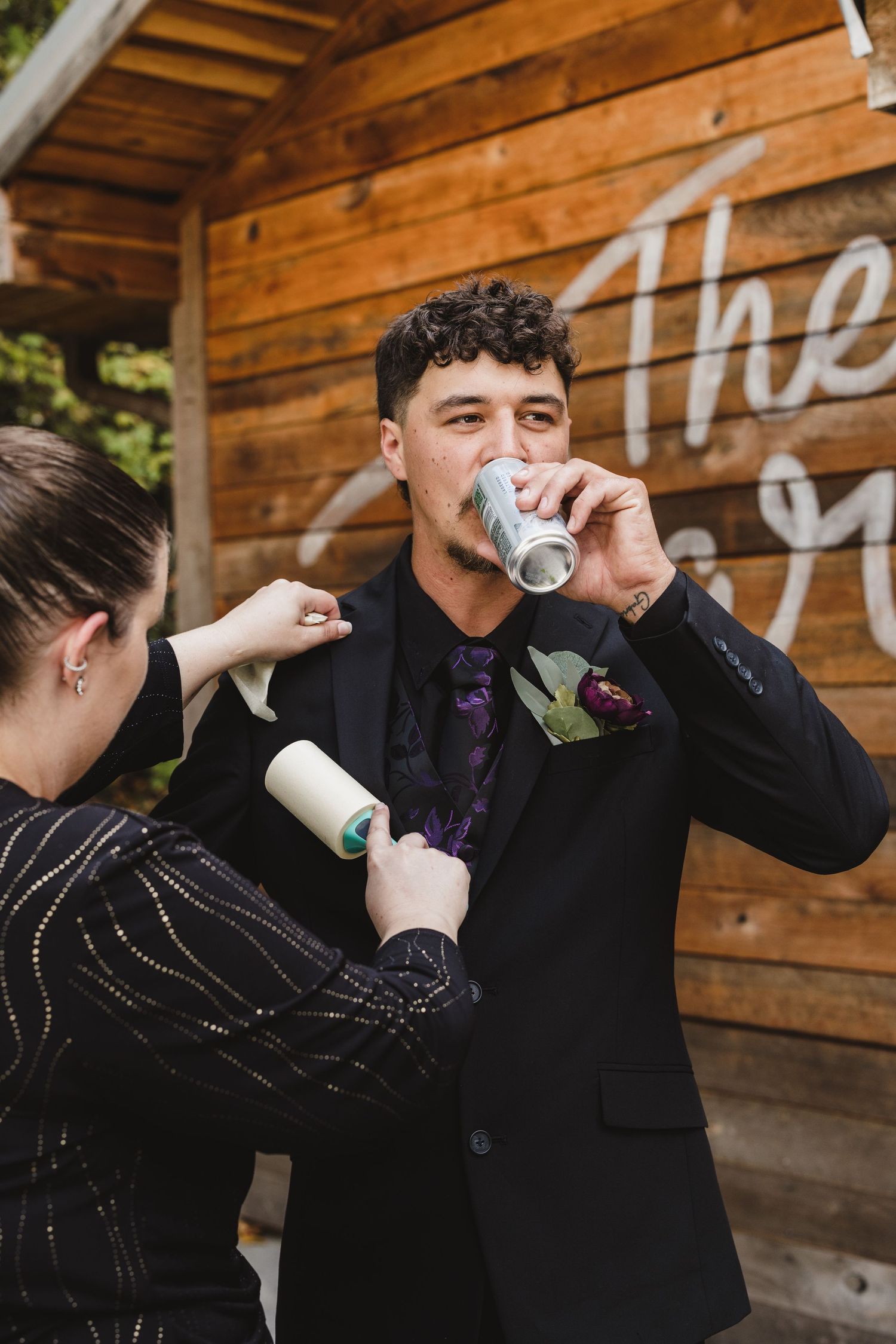 Groom in a black suit with a purple tie and floral boutonniere drinks from a can while a woman uses a lint roller on his jacket.