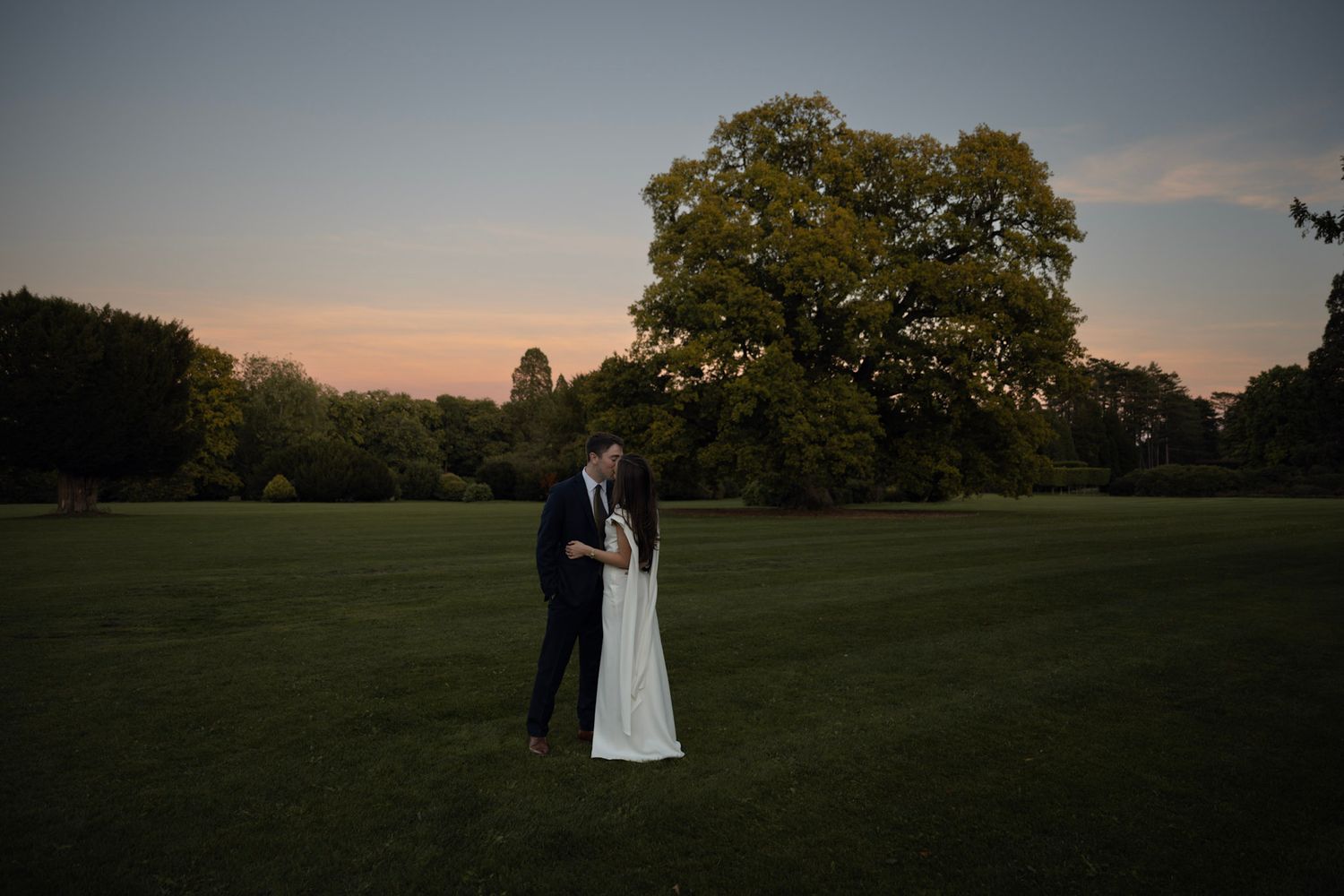 A couple stands together on a grassy field at sunset with a large tree in the background.