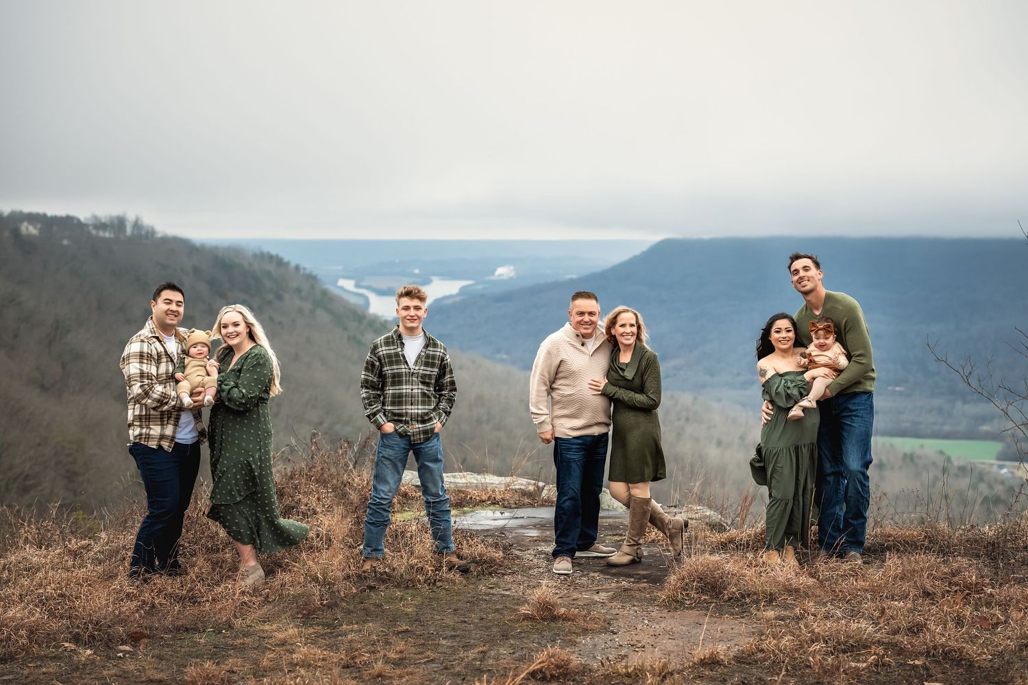 An extended family stand together outdoors on a hilltop overlooking a scenic mountain landscape in winter.