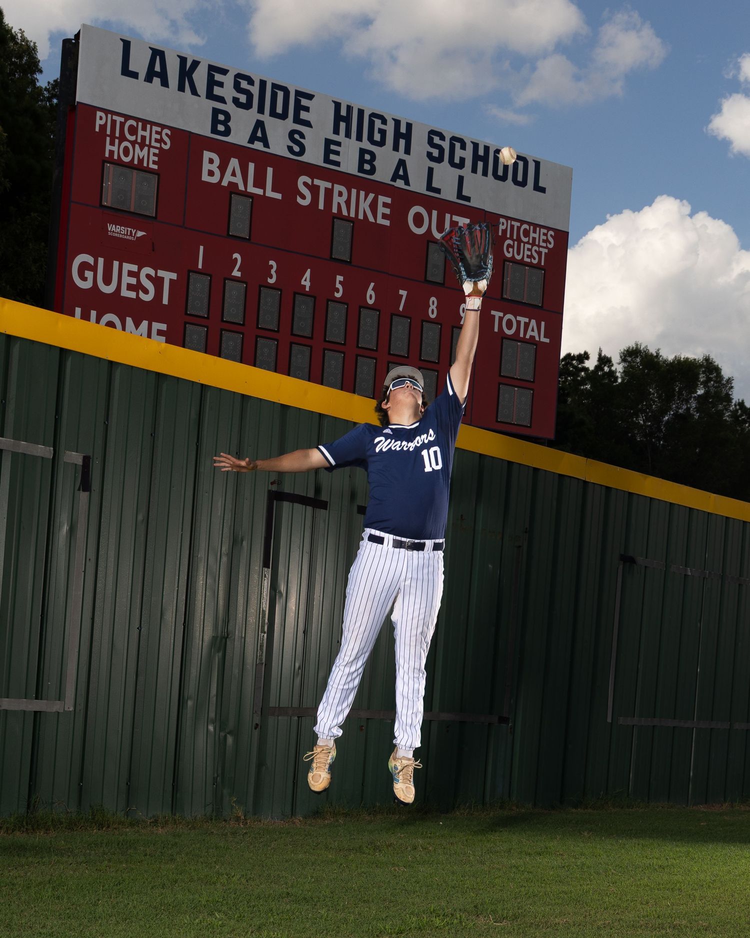 Baseball player swings bat at dusk on dirt field with dramatic cloudy sky and sun rays in background.