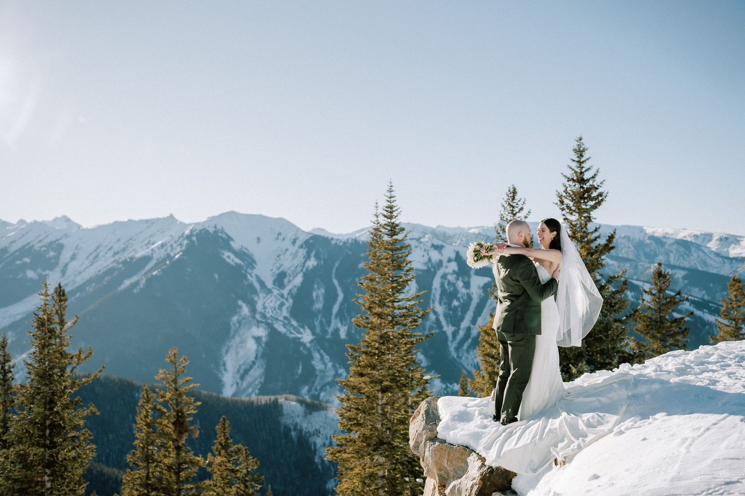 A couple celebrates their wedding surrounded by snowy ground and majestic mountains at the top of Aspen Mountain. Elopement photography.