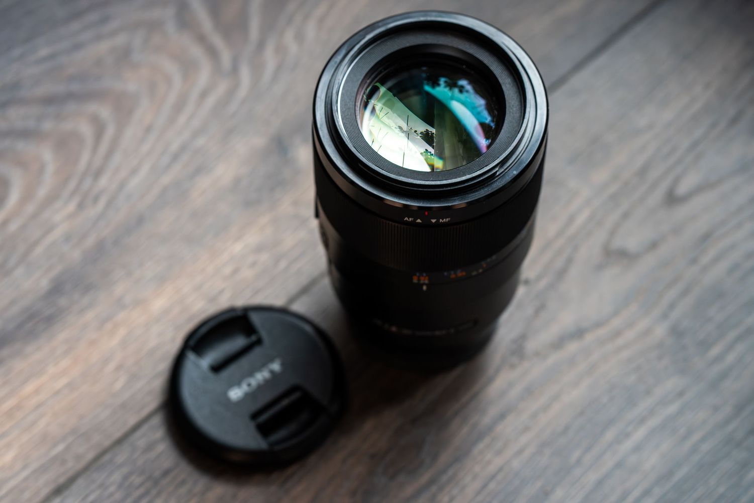 A Sony camera lens with lens cap sits on a wooden surface with visible wood grain pattern.