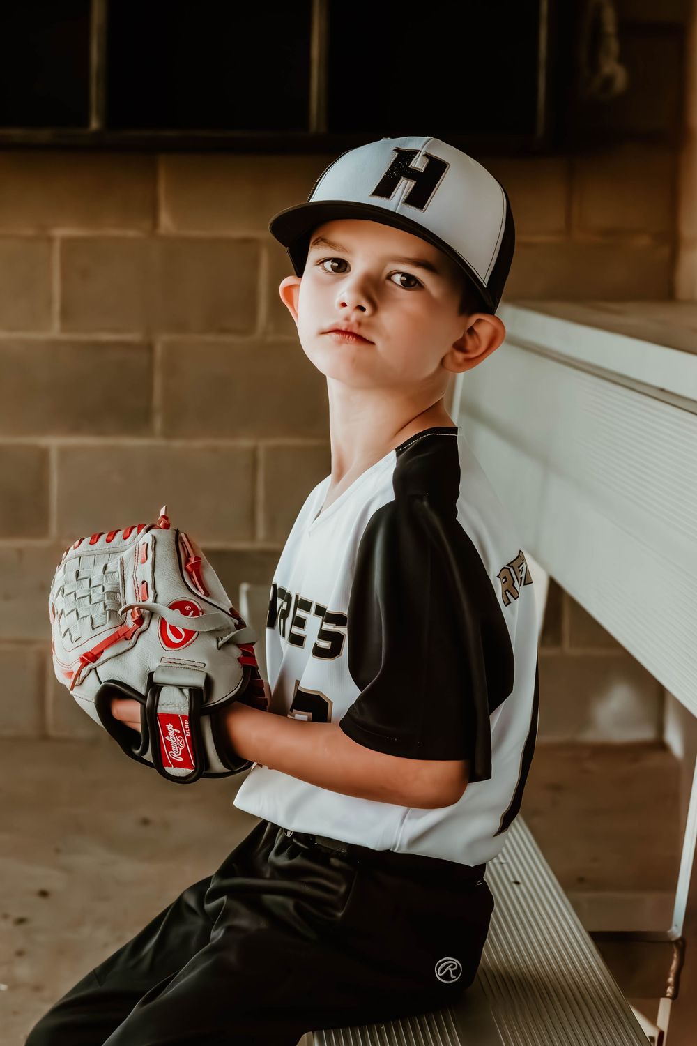 Young baseball player in uniform poses with baseball glove in dugout.