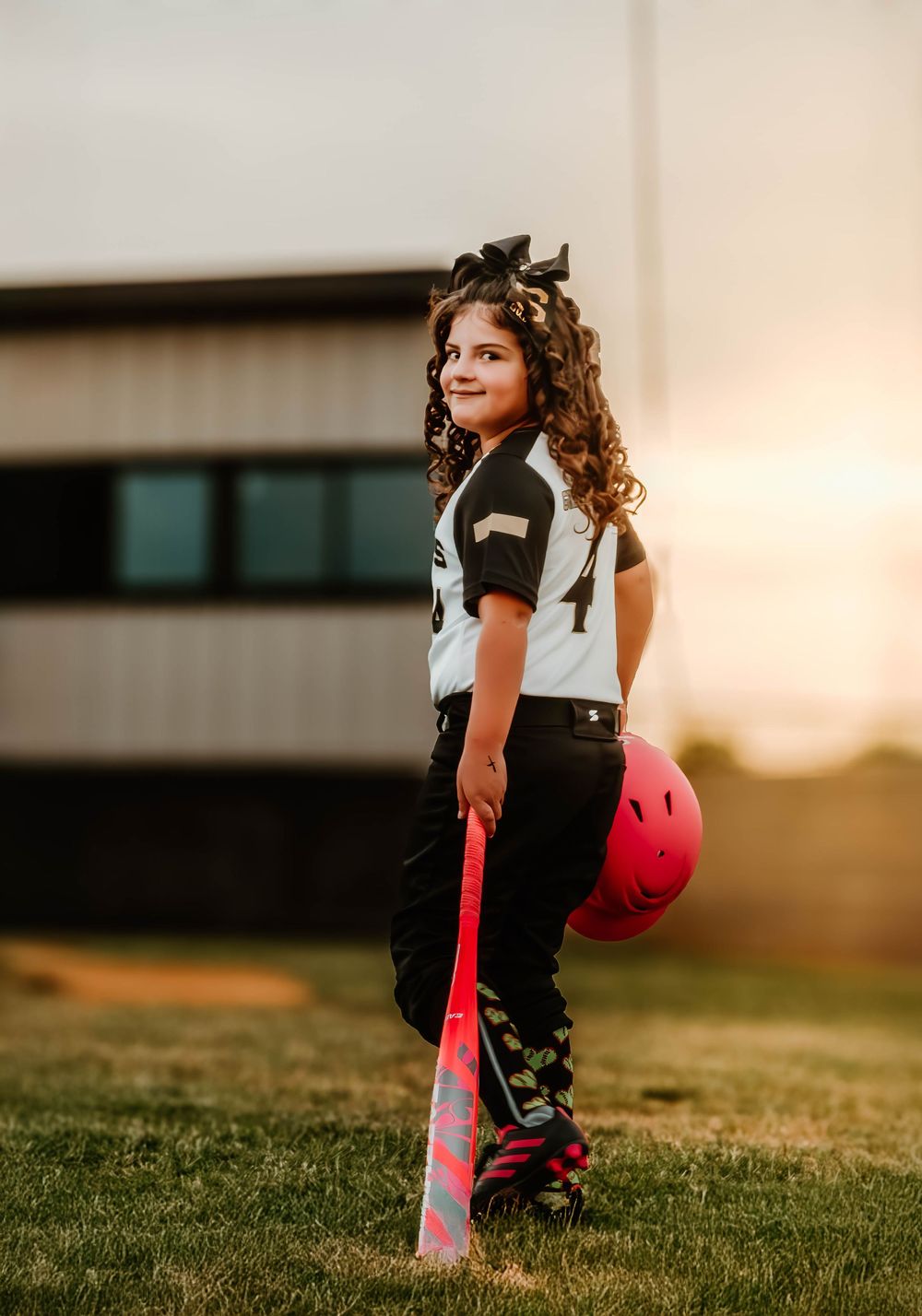 Softball player stands in grass field holding pink bat during golden hour.
