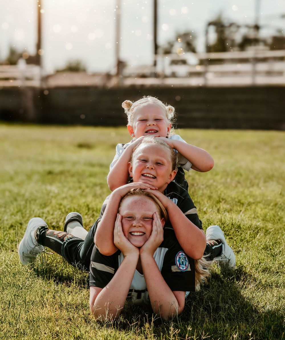 Three young softball players pose playfully on grass in their uniforms.