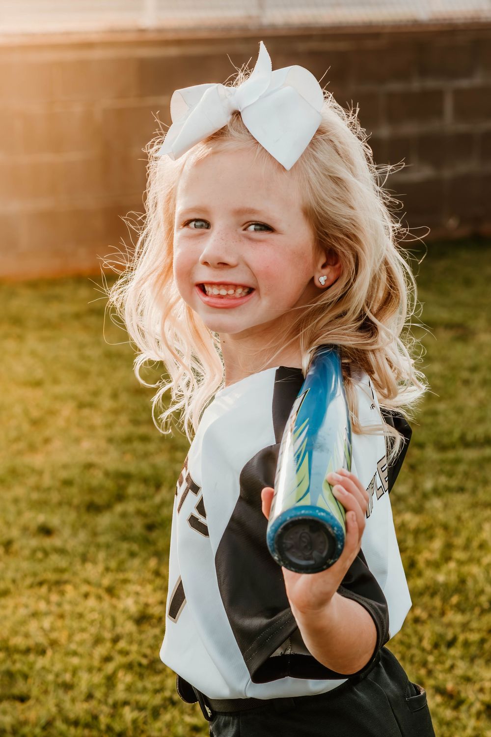 Young softball player in uniform and bow smiles while holding blue water bottle.