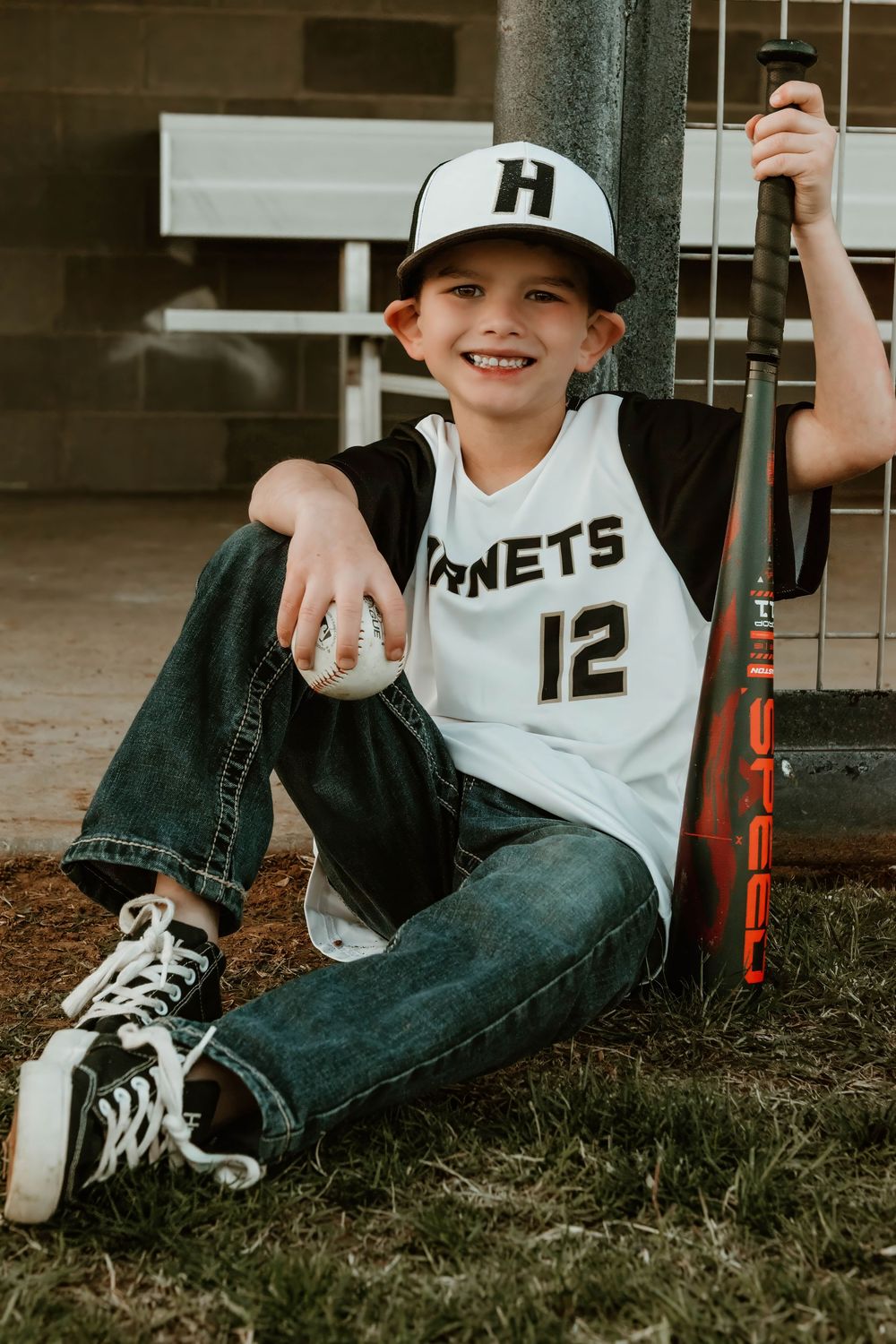 Youth baseball player in uniform number 12 sits on grass near field.