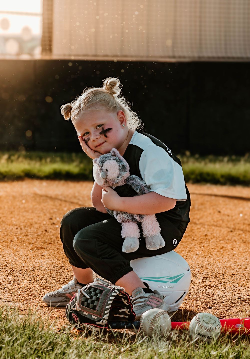 Young softball player in uniform sits with equipment on field during golden hour.