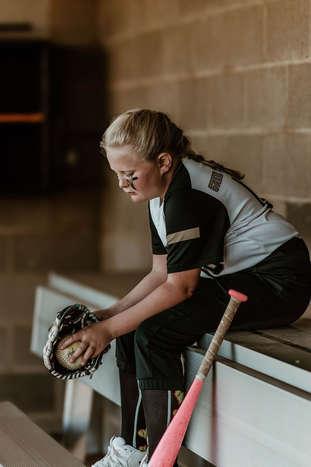 Young softball player in black and white uniform leans over dugout railing.