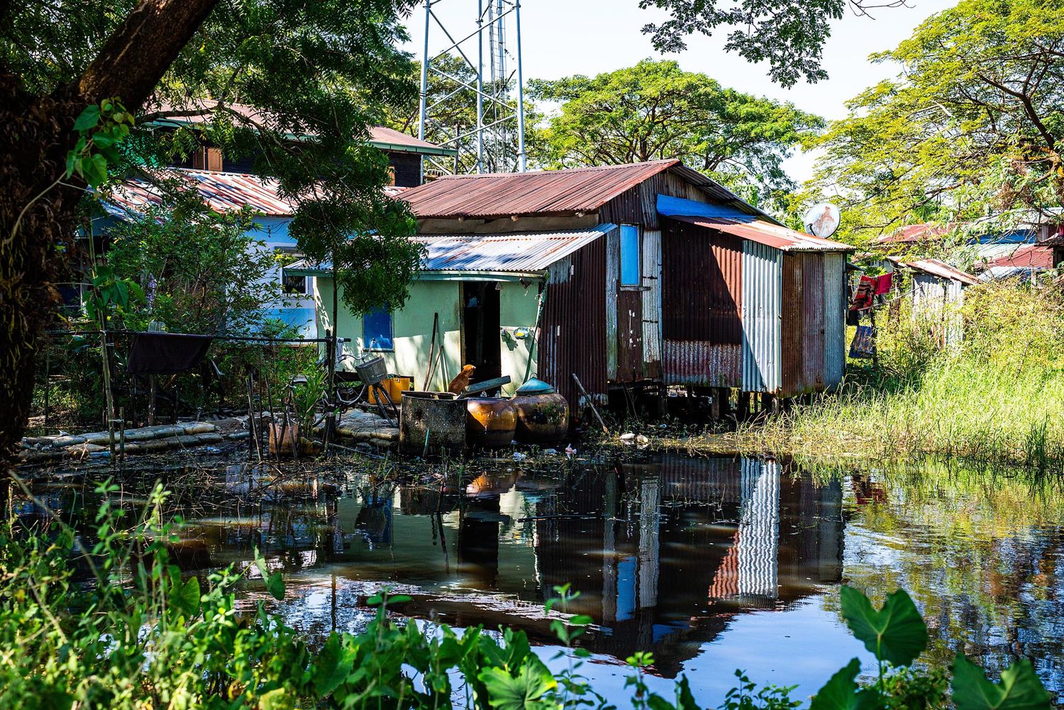 Rustic waterfront houses with tin roofs reflect in still water among tropical vegetation in a rural village setting.