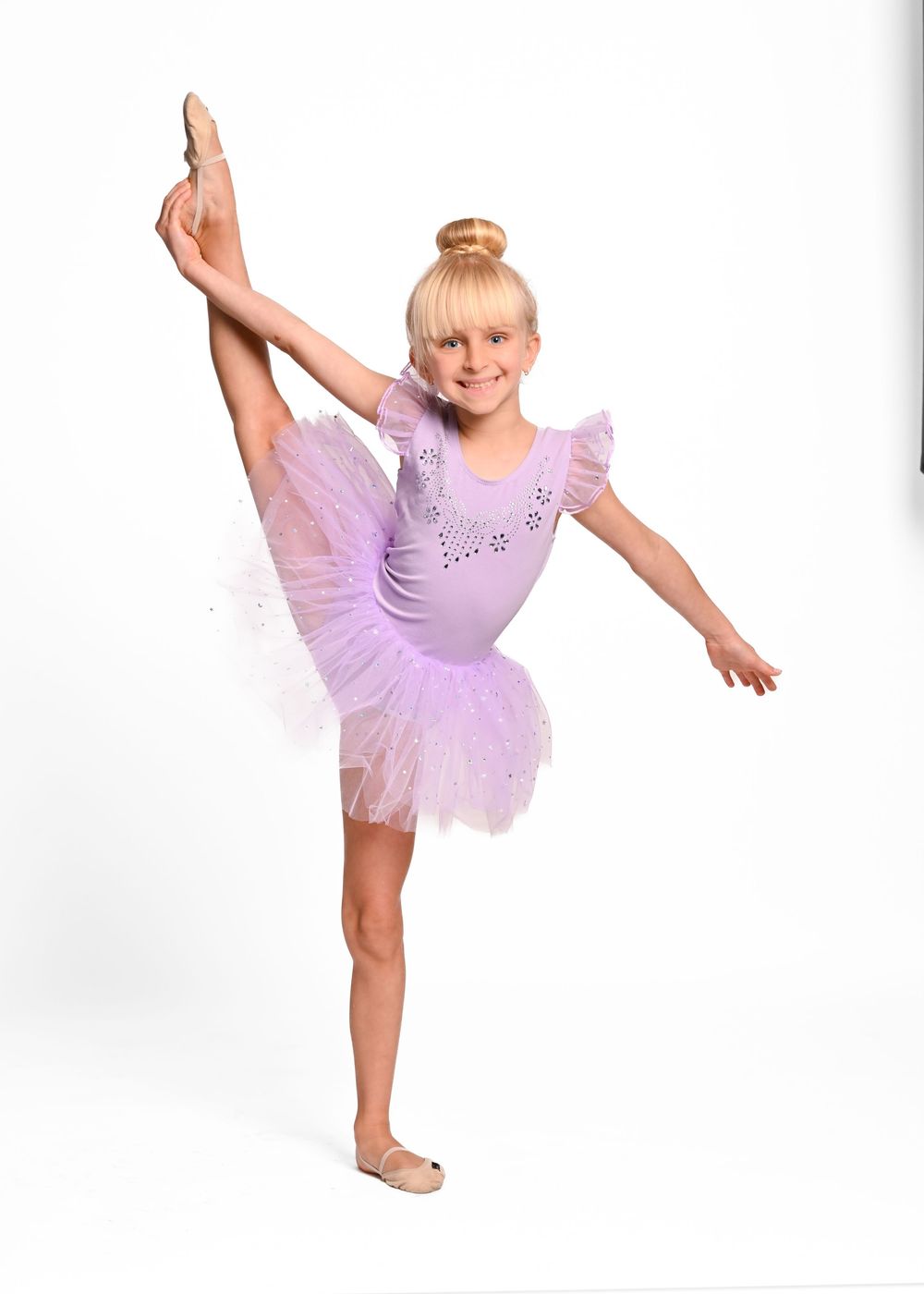 Young dancer in lavender tutu performs graceful ballet pose with extended leg and raised arms.