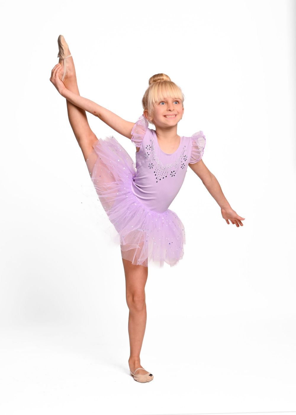 Young ballet dancer in purple leotard and tutu performs graceful leg extension pose against white background.