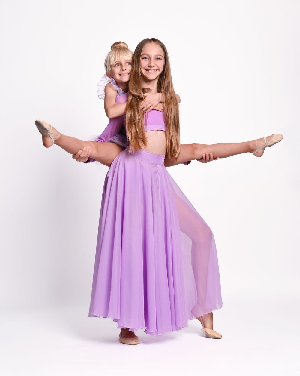 Two dancers in flowing purple dresses perform graceful poses against a white background.