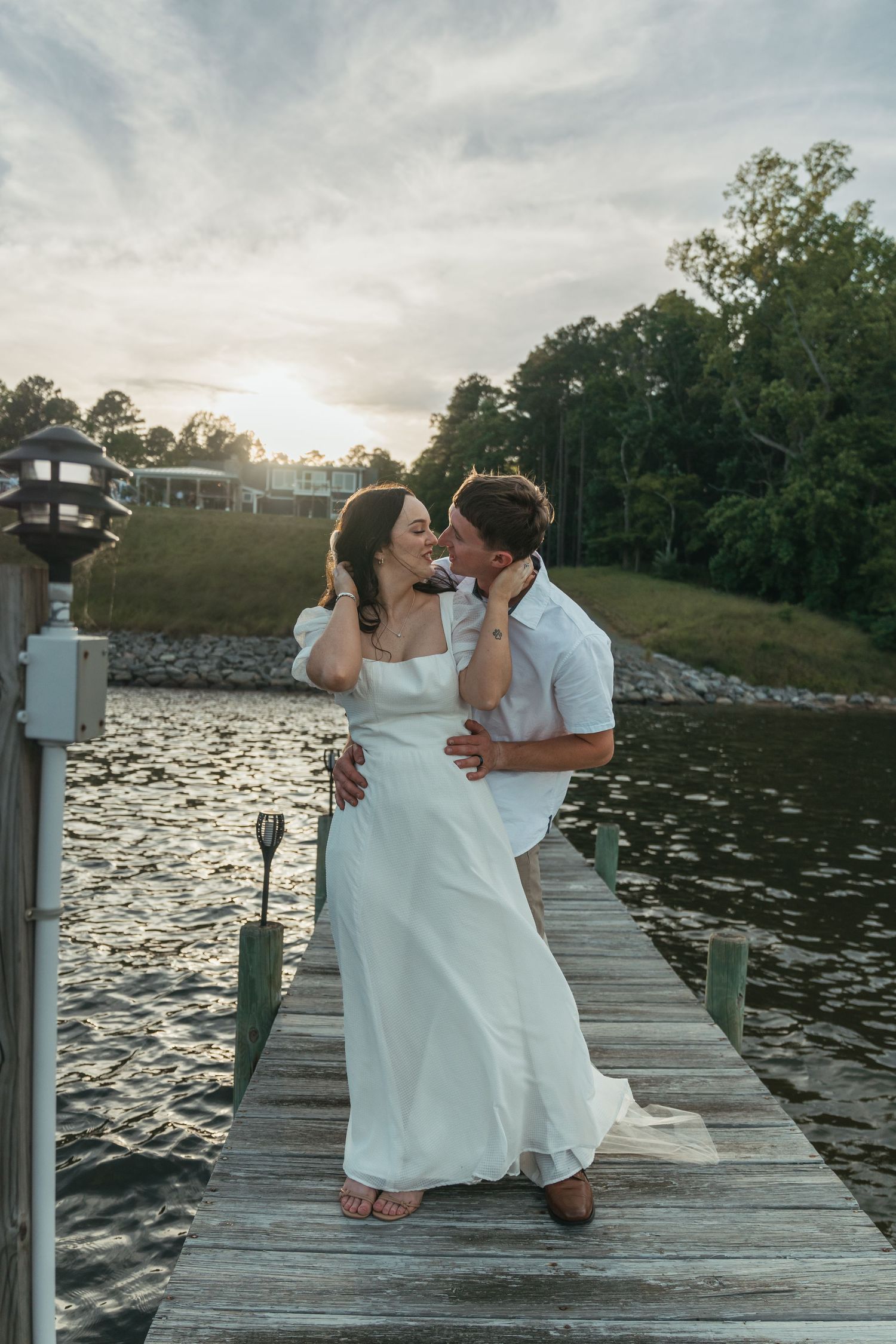 Wedding couple shares romantic moments on wooden dock by lake at sunset.