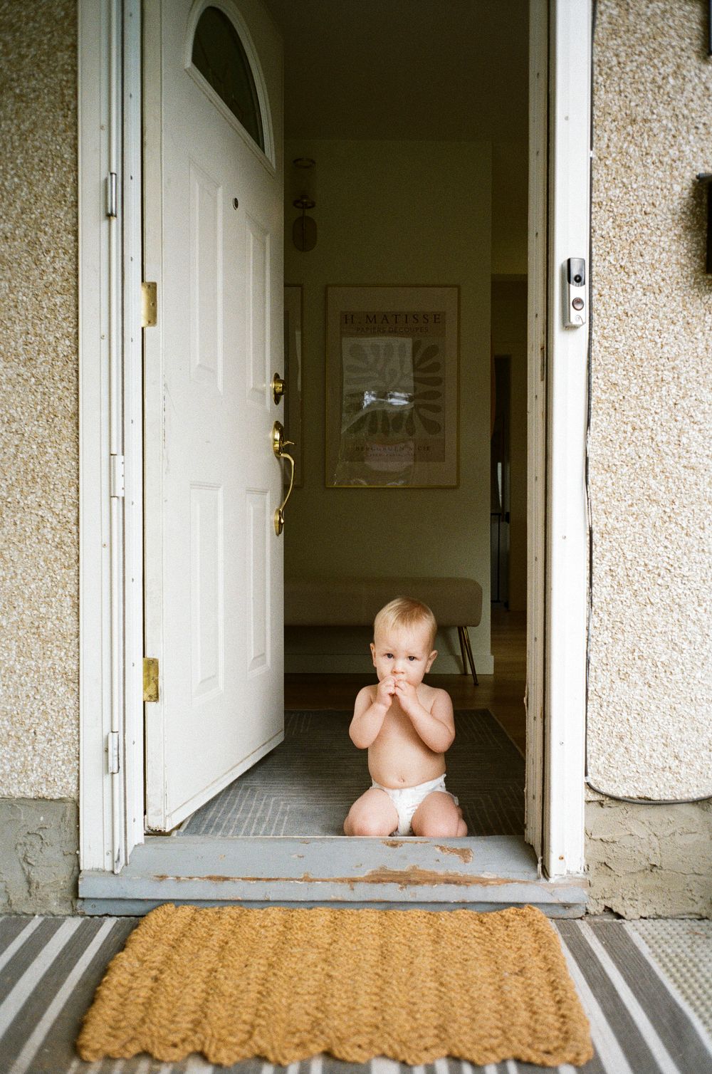 A small child in a diaper sits at a doorway entrance with a welcome mat and white door.
