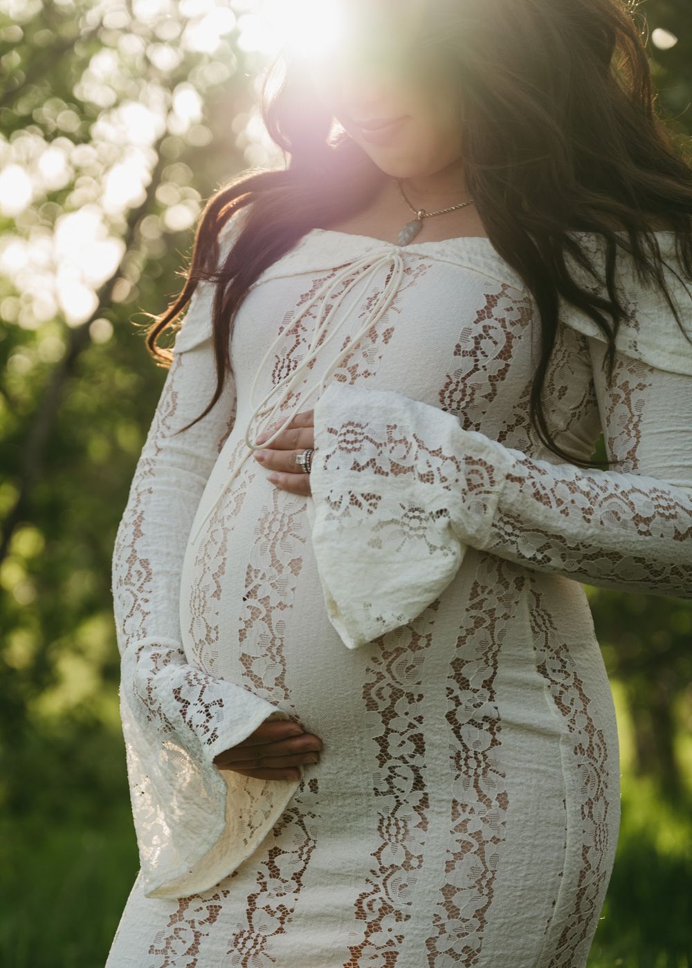 Ethereal maternity photo in white lace dress with sunlight streaming through trees in the background.