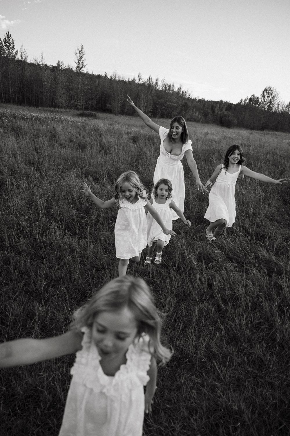 People in white dresses dance and twirl through a grassy field at dusk in black and white photography.
