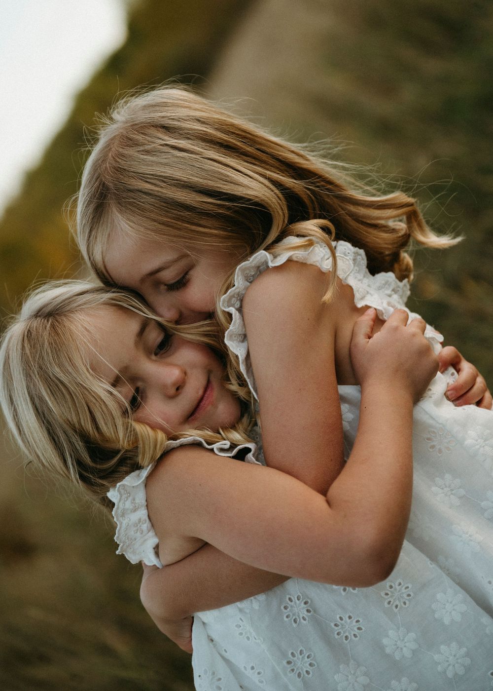 Two children in white dresses share a tender embrace while playing outdoors.