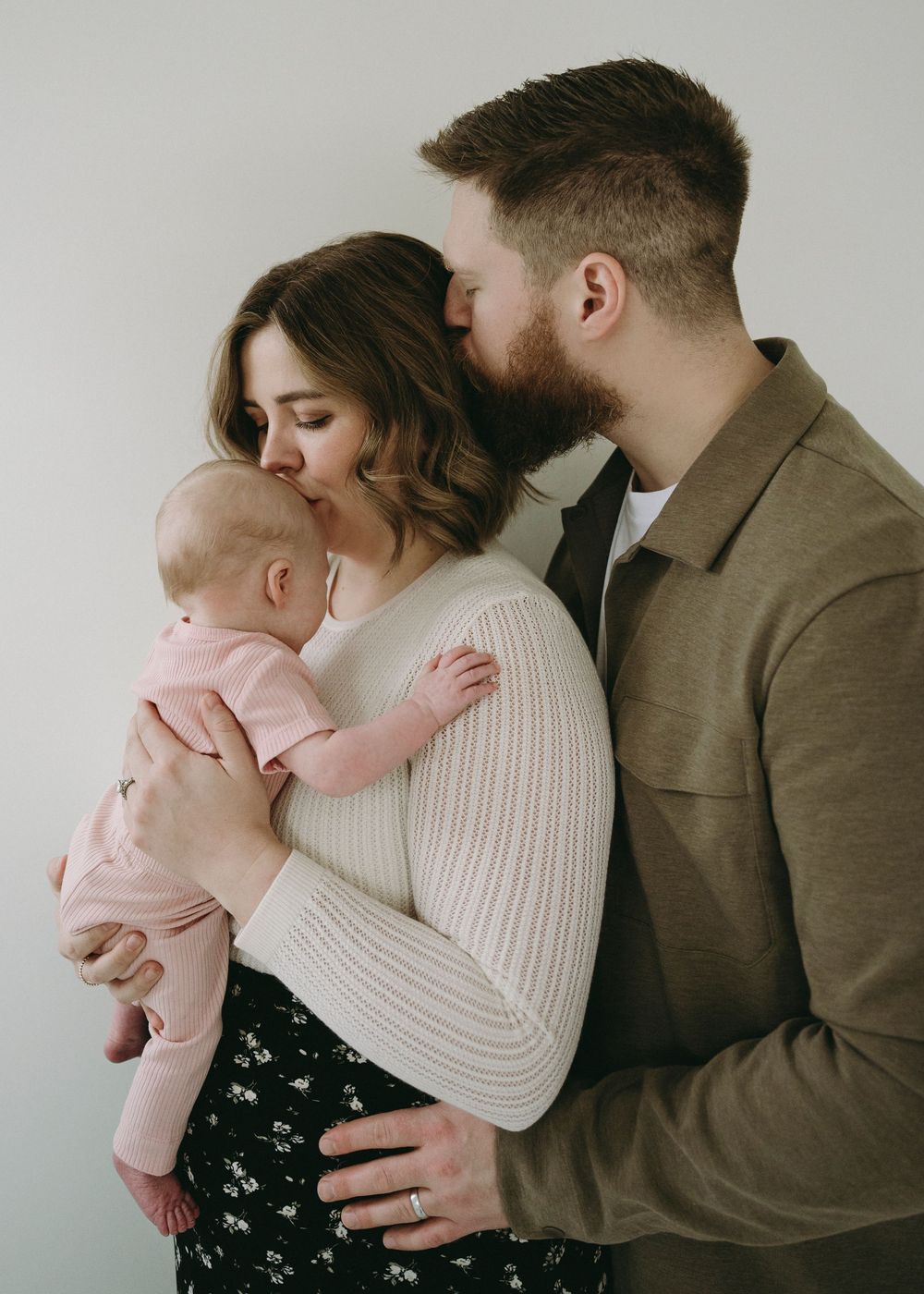 A couple in neutral tones embraces tenderly while holding their baby in pink against a light background.