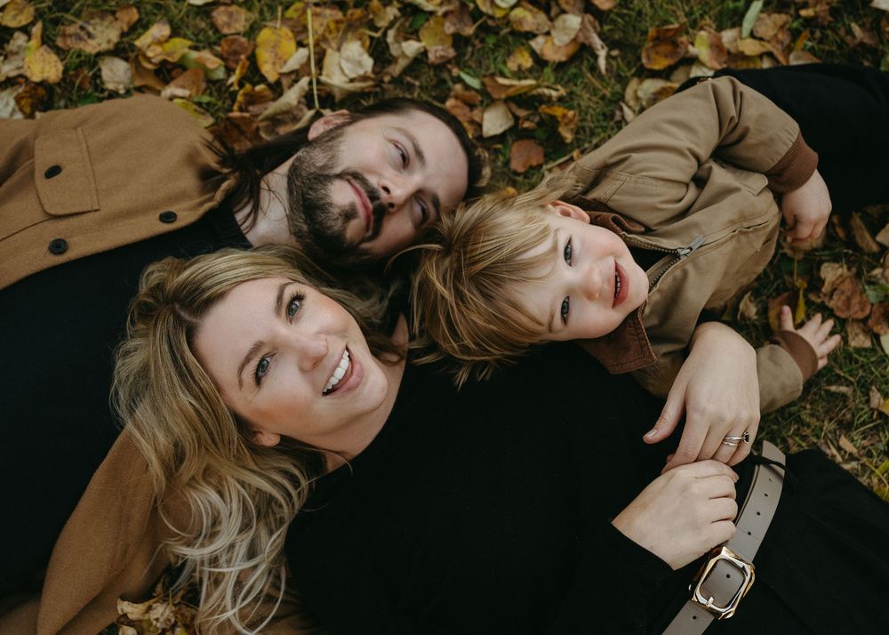 Three people lying on fallen autumn leaves wearing brown and tan outerwear while smiling at the camera.