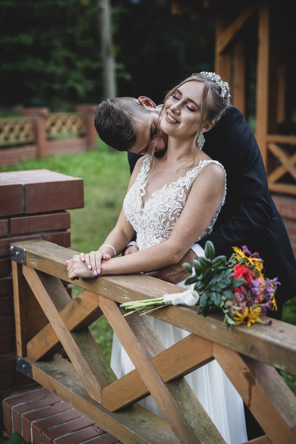 Couple sitting together on rustic wooden bench with colorful wedding bouquet.