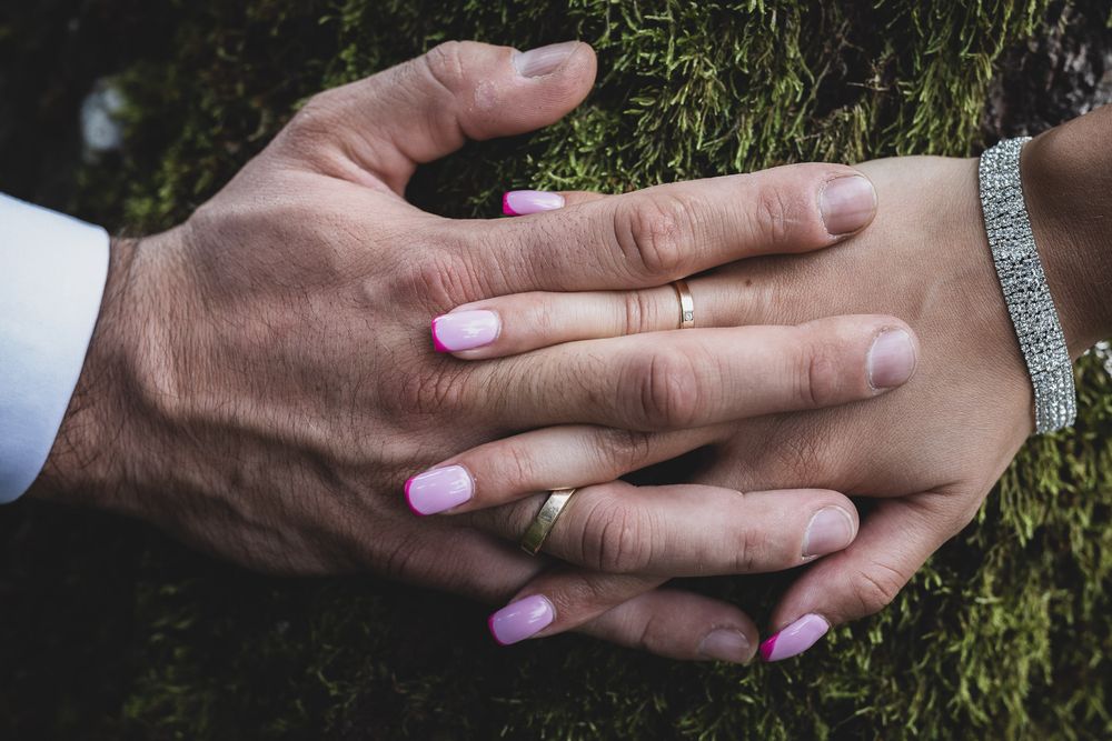 Close up of hands with wedding rings touching on natural green background.