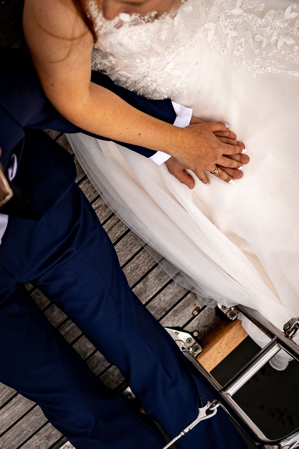 Close-up details of a white wedding dress and navy suit on a wooden dock.