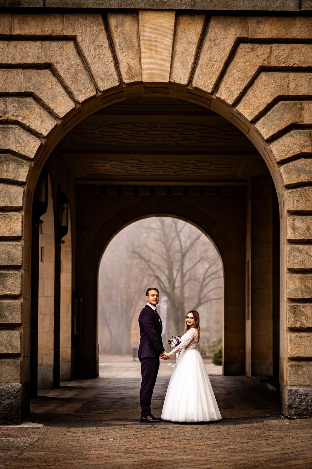 Wedding couple posing under stone archways in dramatic lighting.