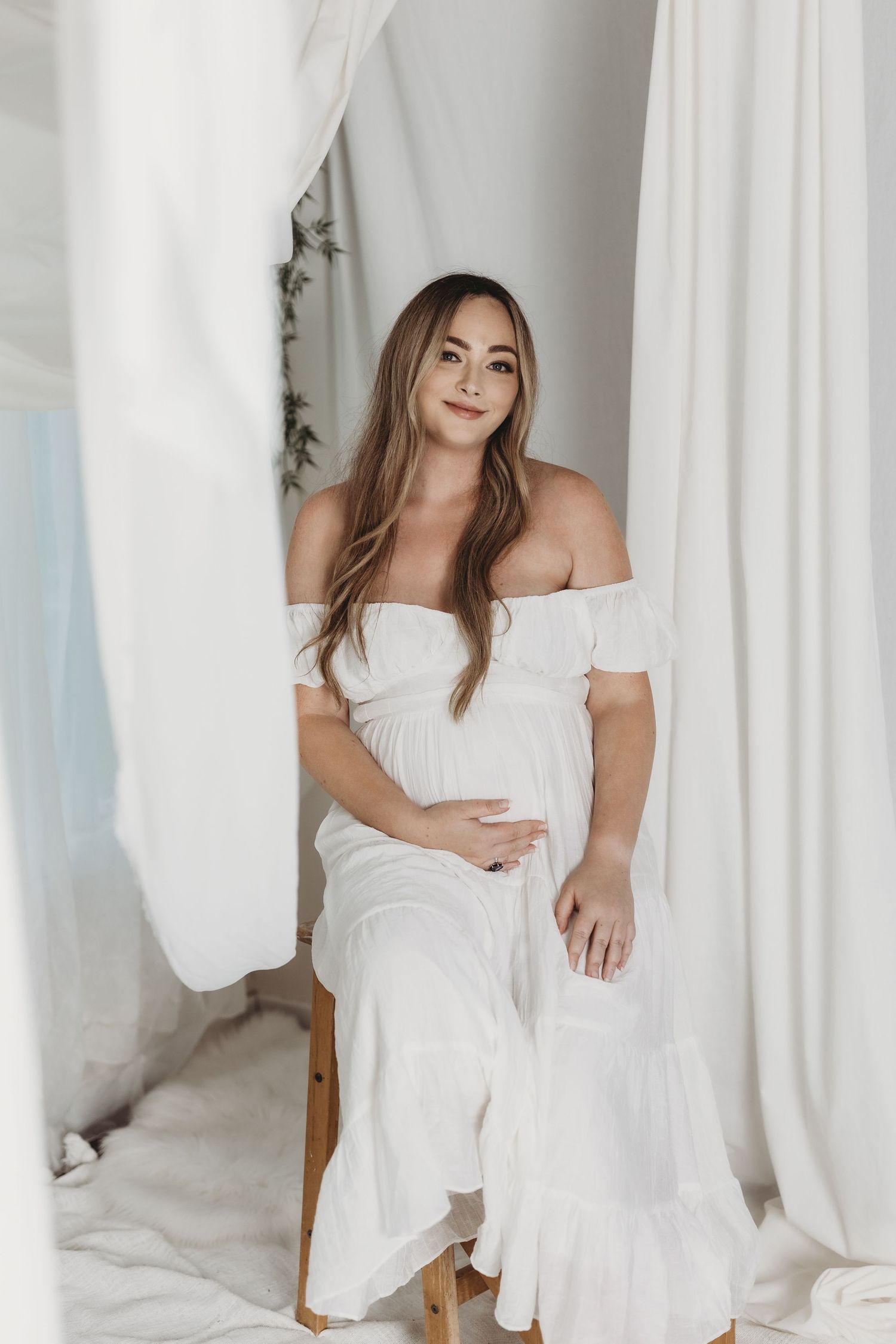 Woman in white dress poses against white curtains in bright interior setting.