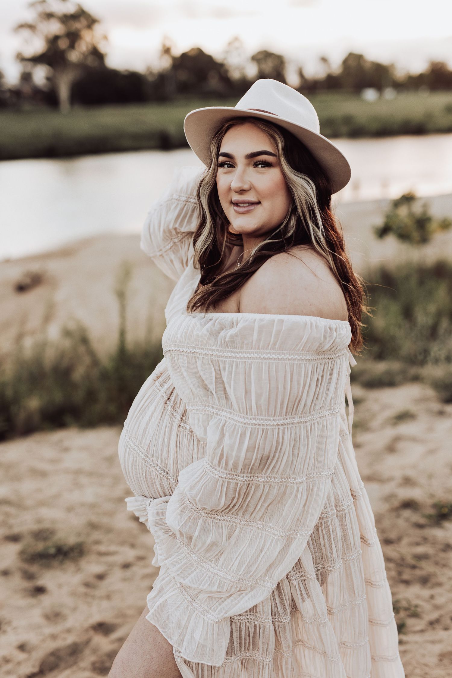Woman in white ruffled dress and hat poses in natural outdoor setting.