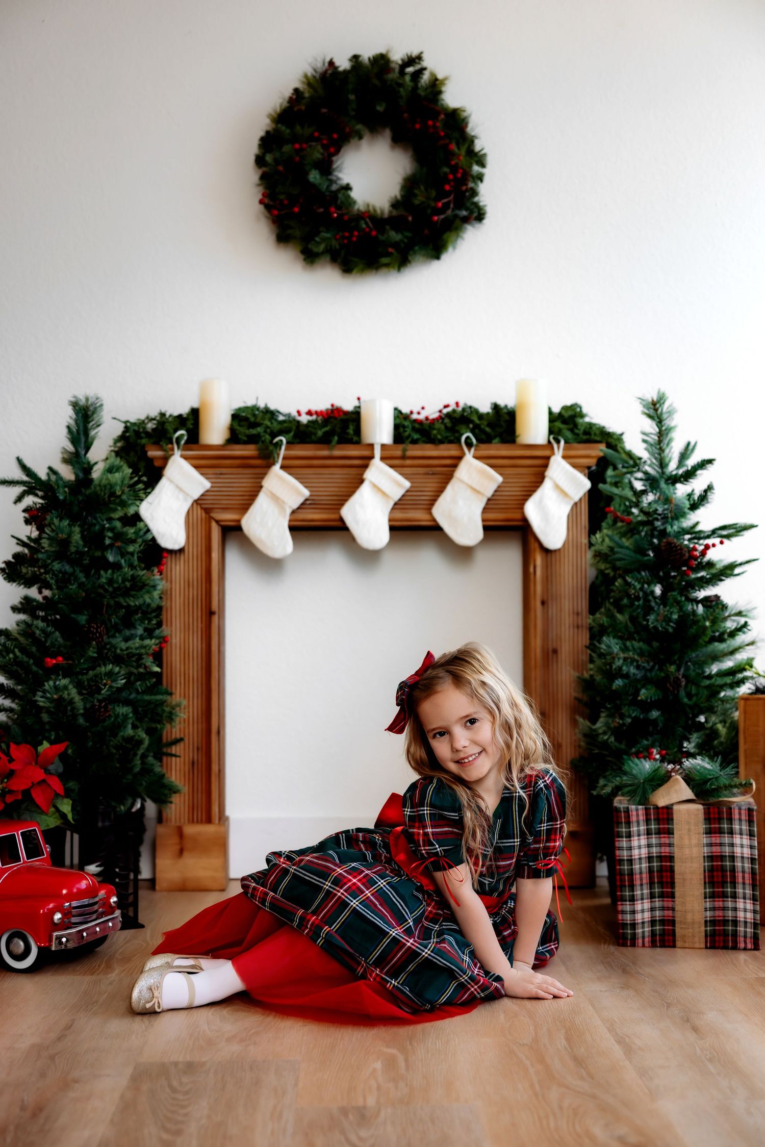 Holiday scene with plaid dress, stockings on wooden mantel, evergreen wreath and wrapped presents on hardwood floor.