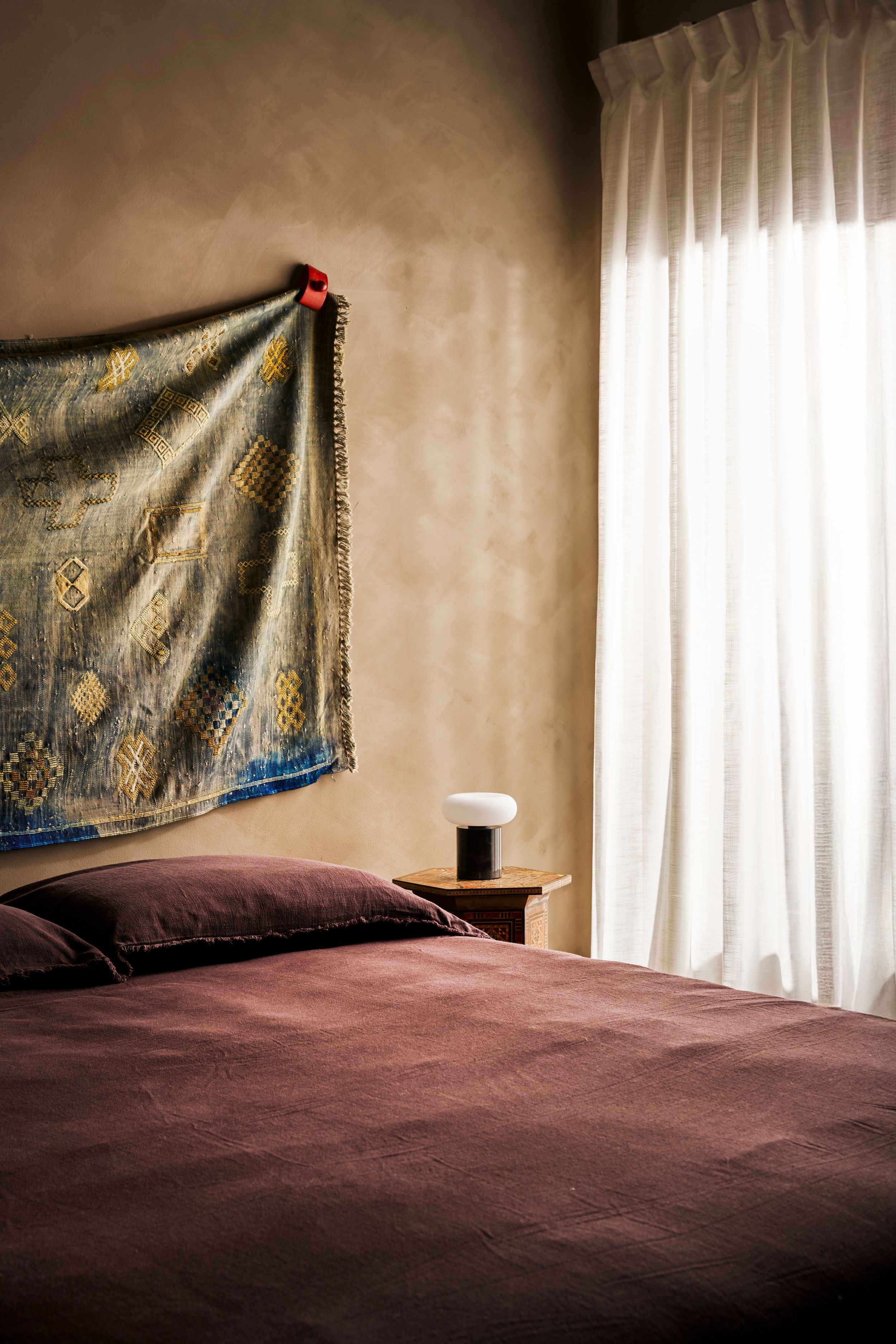 A burgundy bed with a decorative tapestry headboard is illuminated by soft natural light through sheer curtains.