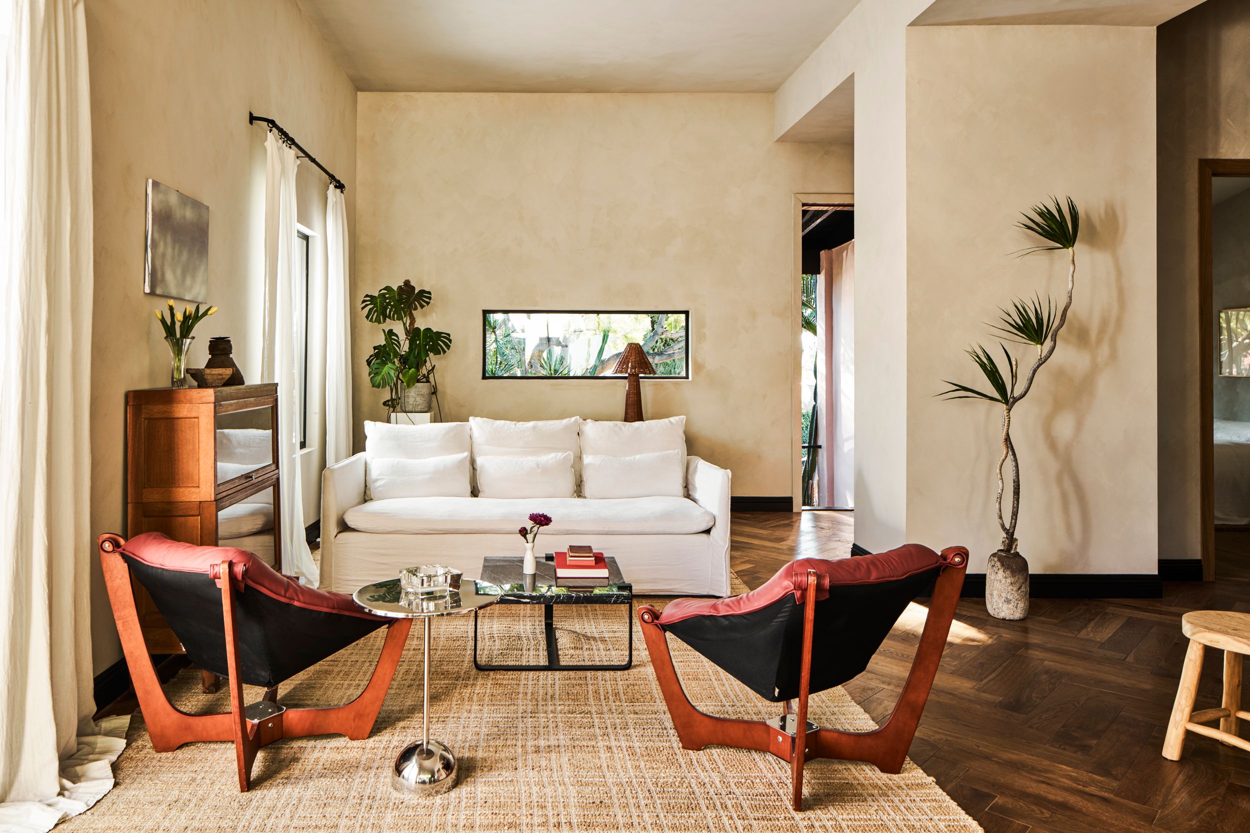 Modern living room interior with coral accent chairs, houseplants, and natural light streaming through large windows.