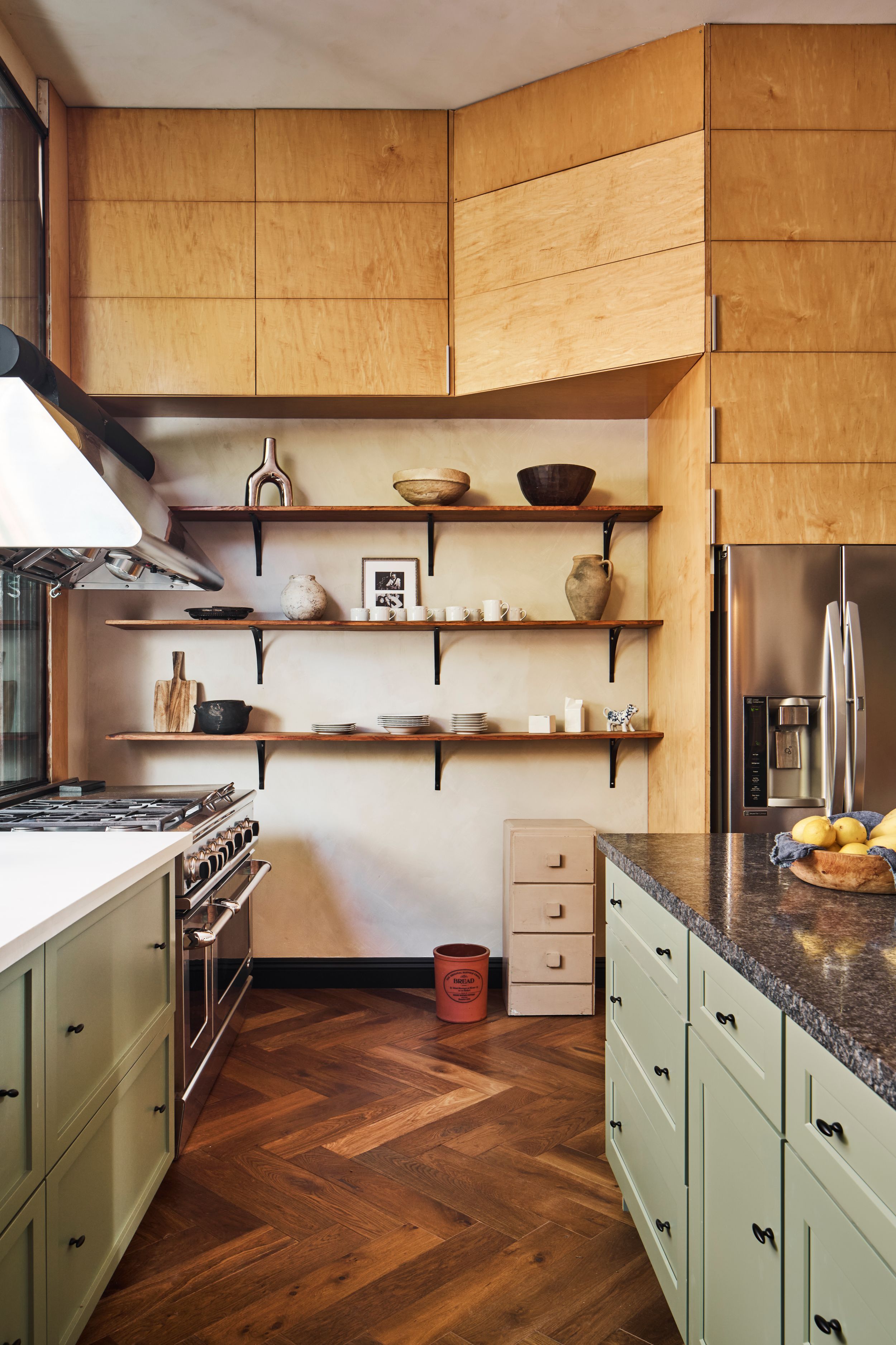 Modern kitchen features sage green base cabinets, wooden upper cabinets, and a dark herringbone wood floor pattern.