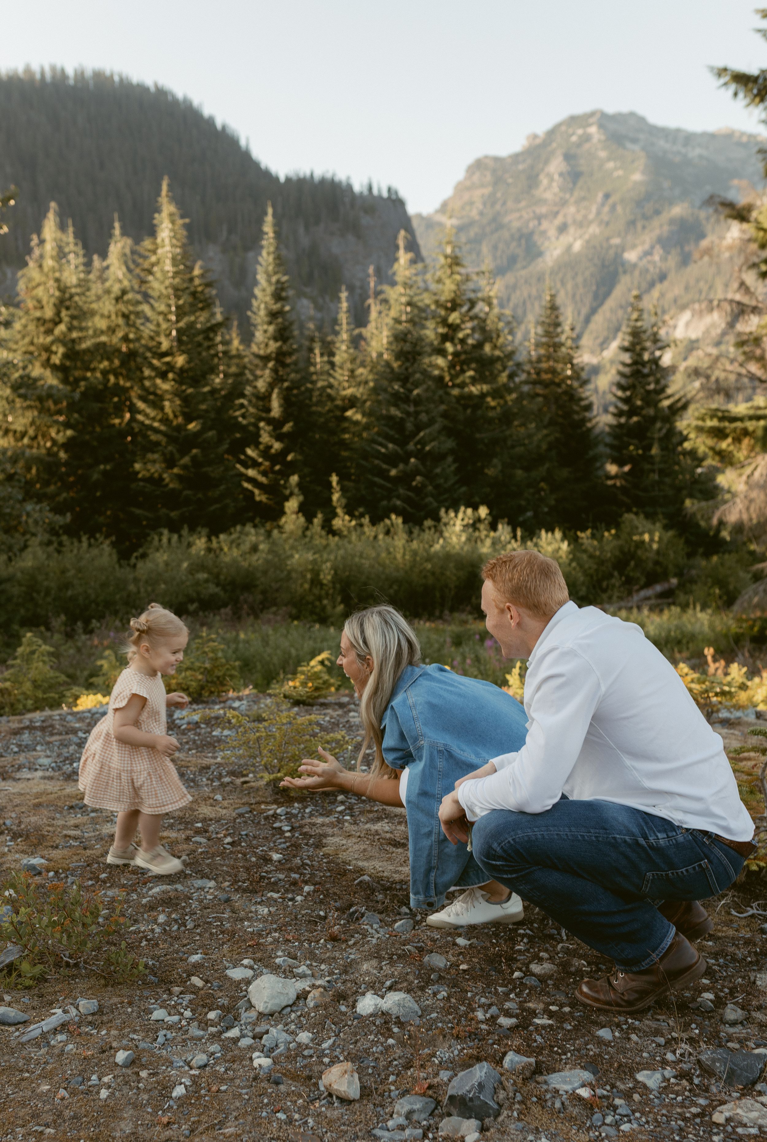 Couple lying on rocky mountain terrain during golden hour with evergreen trees and mountain peaks in background.