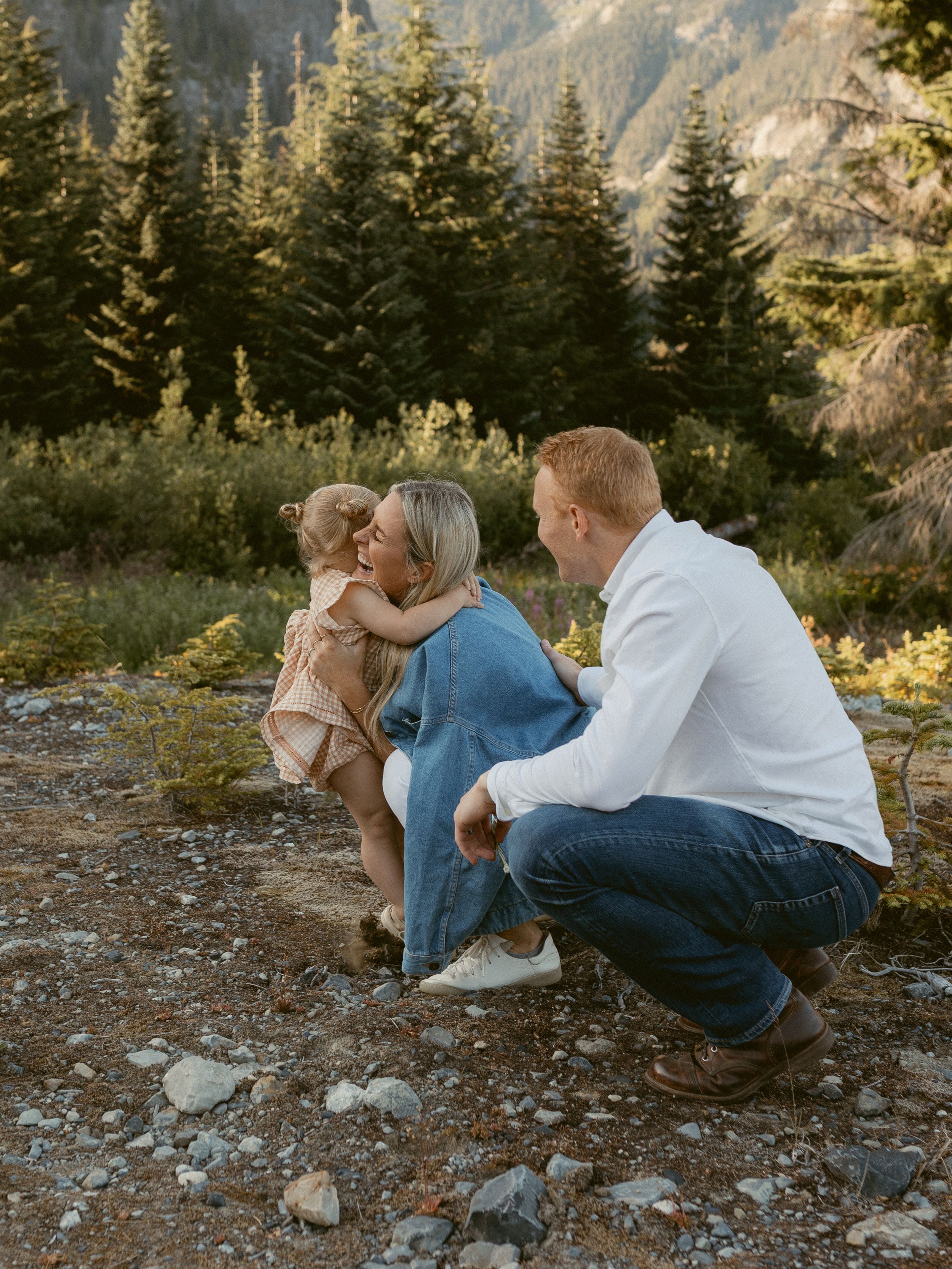 A couple shares an intimate moment while sitting on rocky mountain terrain surrounded by evergreen trees at sunset.