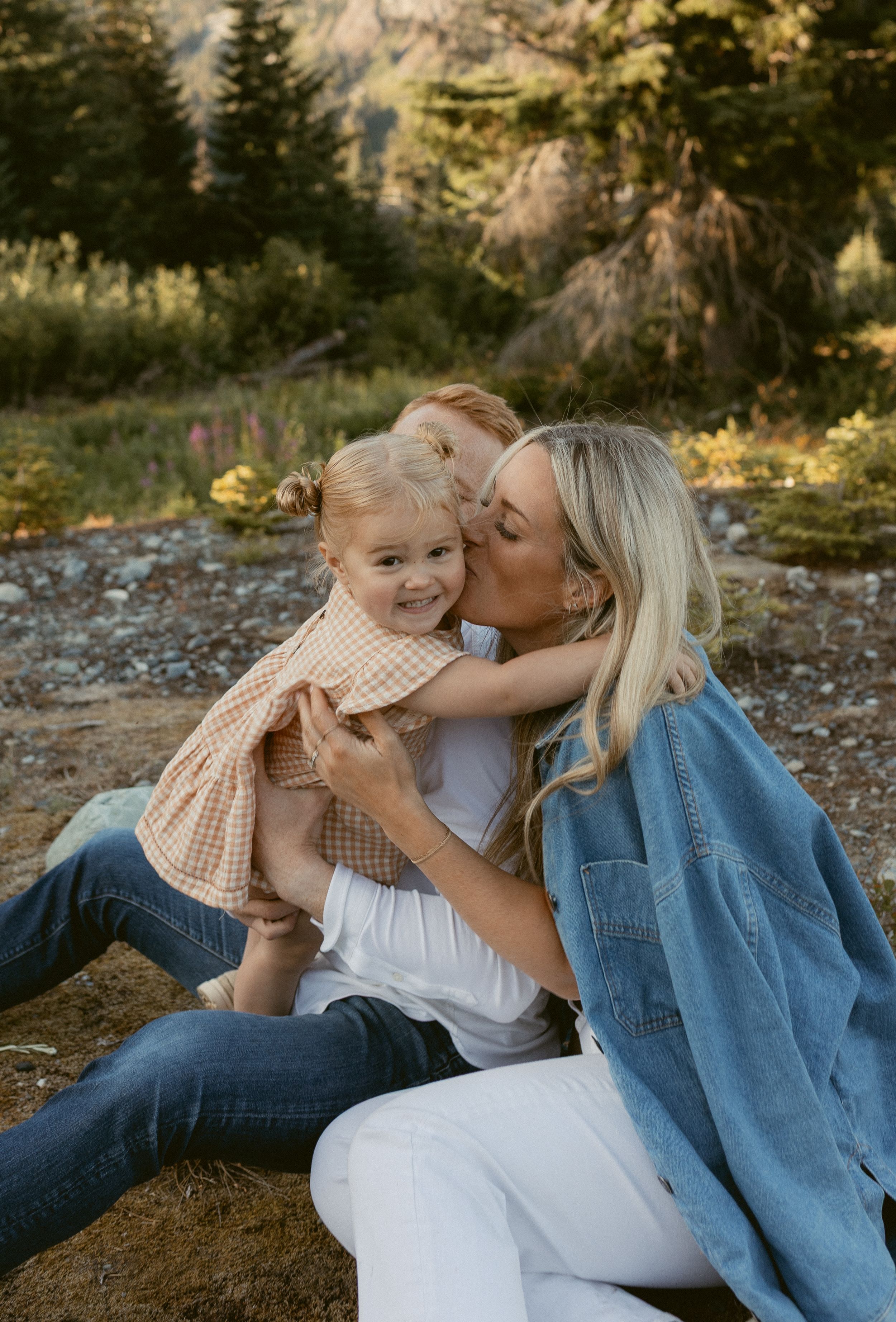 Sweet family moment captured against mountain backdrop with warm golden light filtering through trees.
