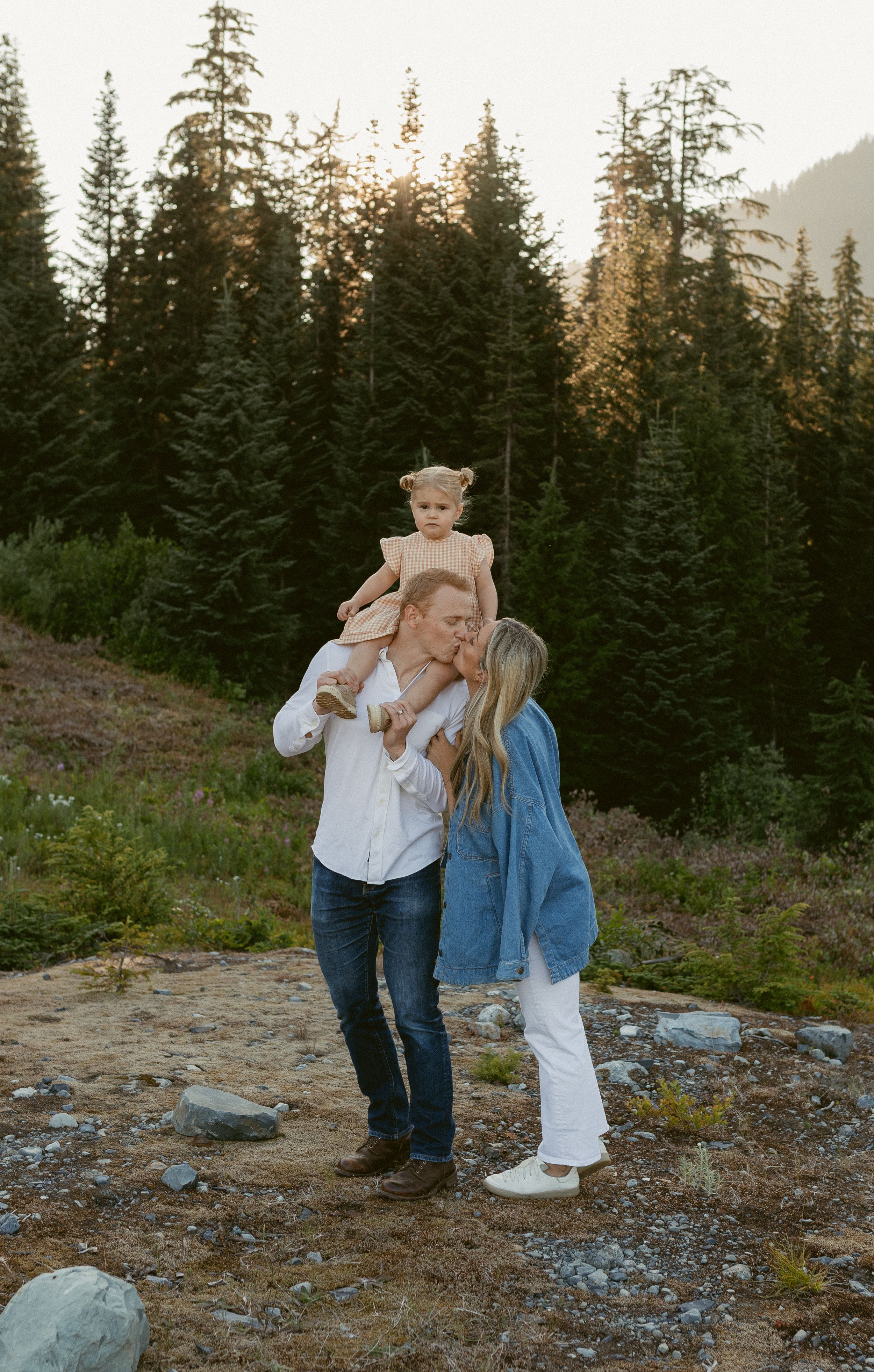 A couple shares a playful moment together on a rocky mountain trail surrounded by evergreen trees at sunset.