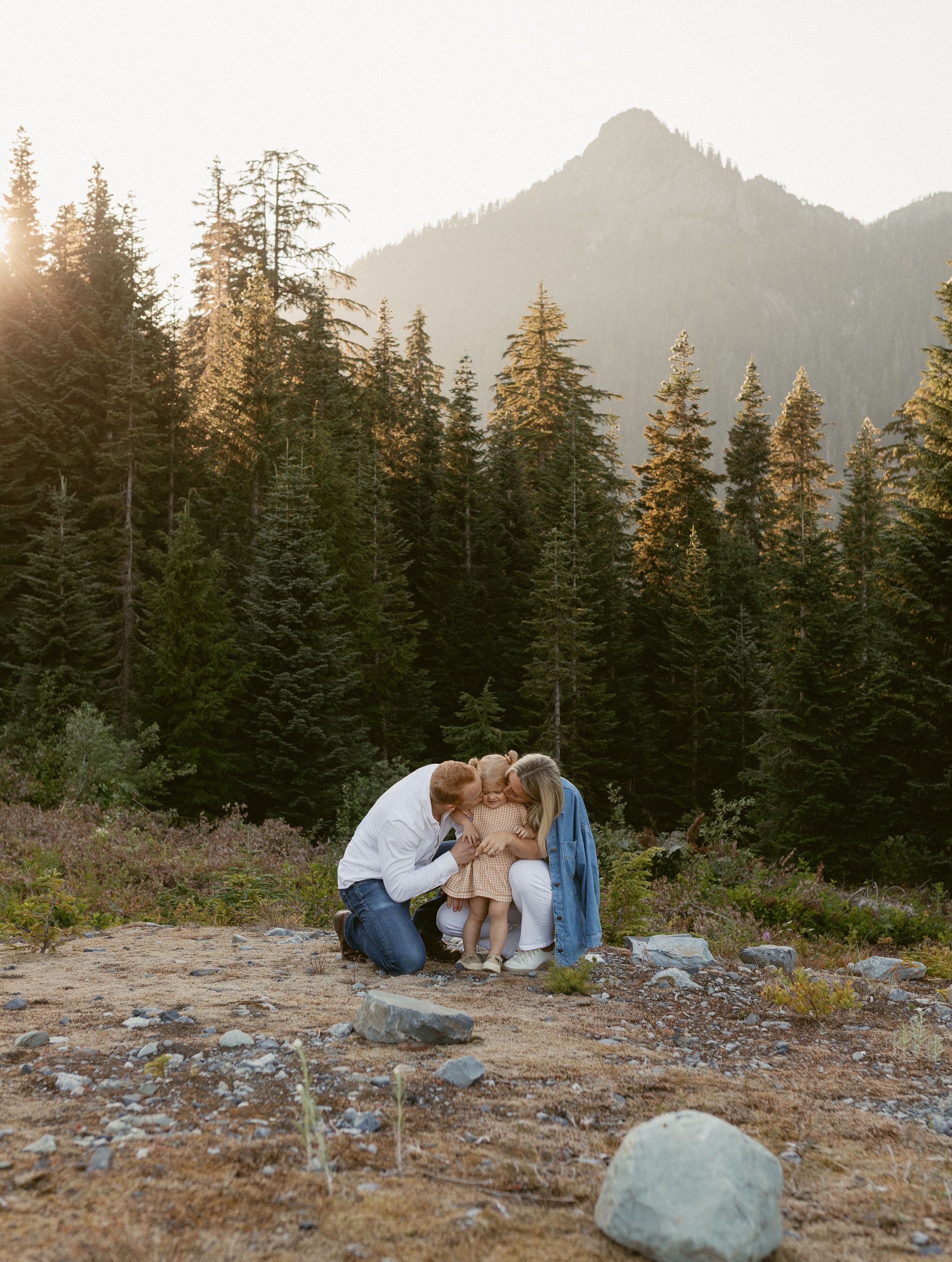 Couple embracing on rocky mountain terrain at sunset with evergreen forest and misty peaks in background.
