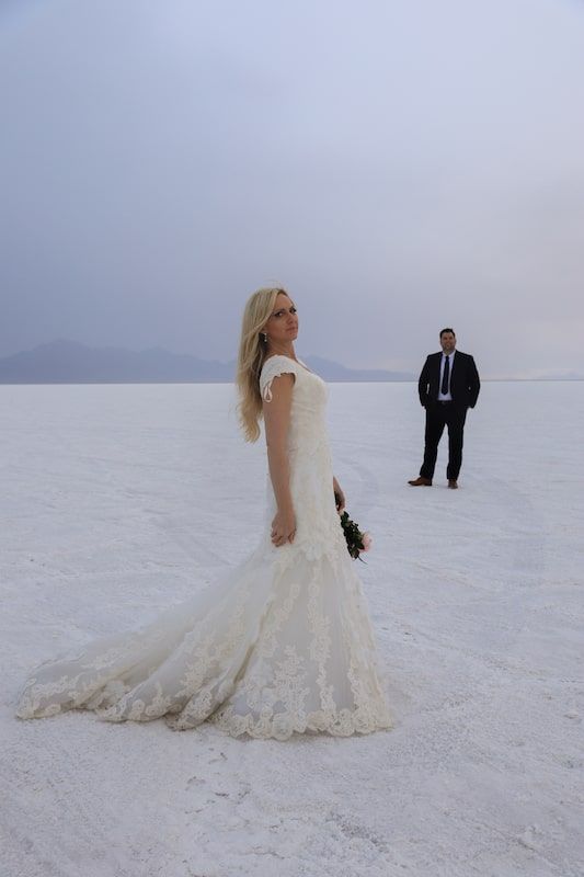 A couple in wedding attire poses on a stark white salt flat landscape under an overcast sky.