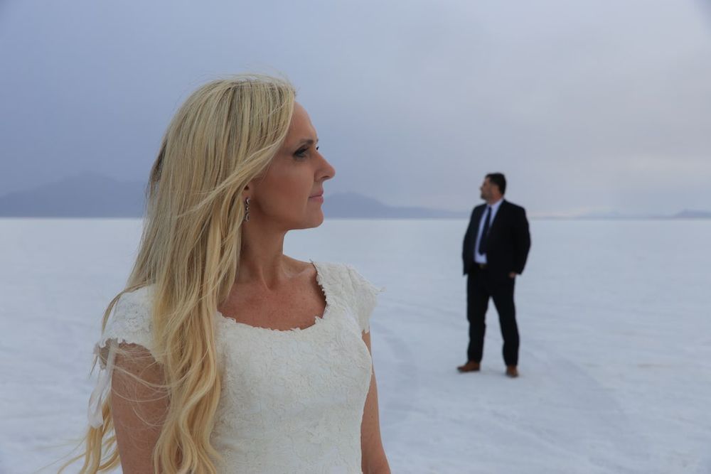 A person with long blonde hair stands in a snowy winter landscape wearing a white dress against a cloudy sky.