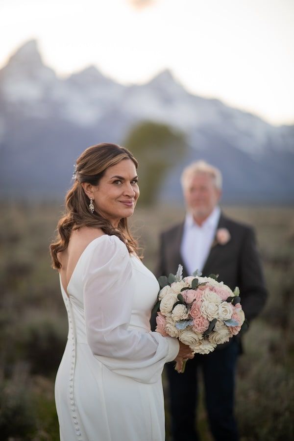 Bride in long-sleeved white wedding dress holds pink and white bouquet against mountain backdrop at sunset.