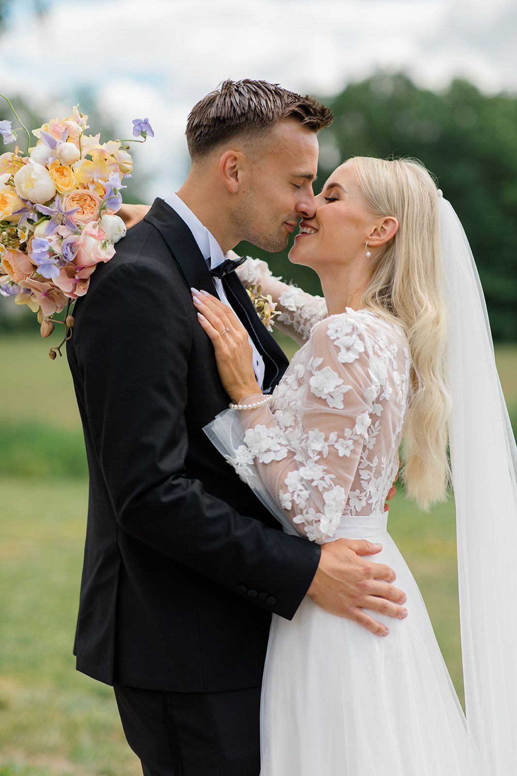 A romantic wedding moment outdoors with a colorful bouquet of peach and purple flowers, lace dress details and classic black tuxedo.