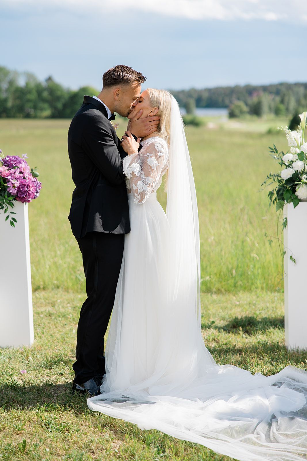 Bride and groom share romantic kiss during outdoor wedding ceremony with floral arrangements and rolling hills in background.