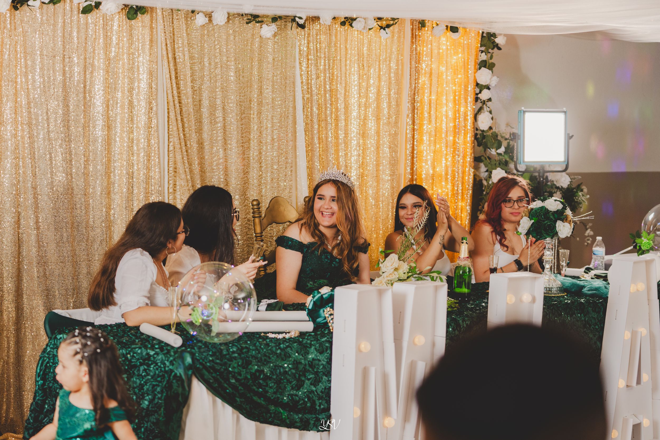 Wedding guests dancing and celebrating at an elegant reception with gold curtains and decorative lighting.
