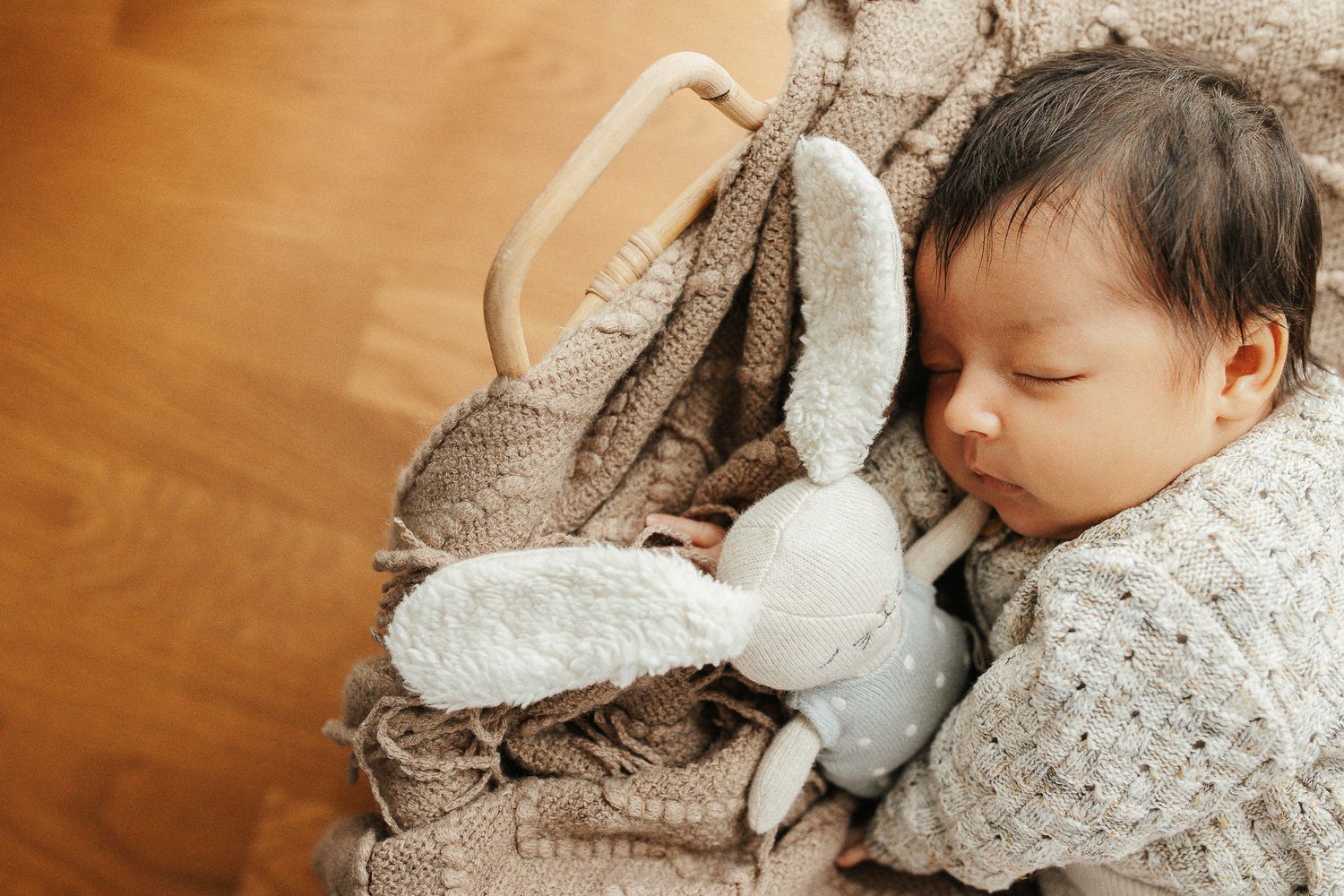 Sleeping baby cuddled with soft plush bunny toy on wooden floor with cozy knit blanket.