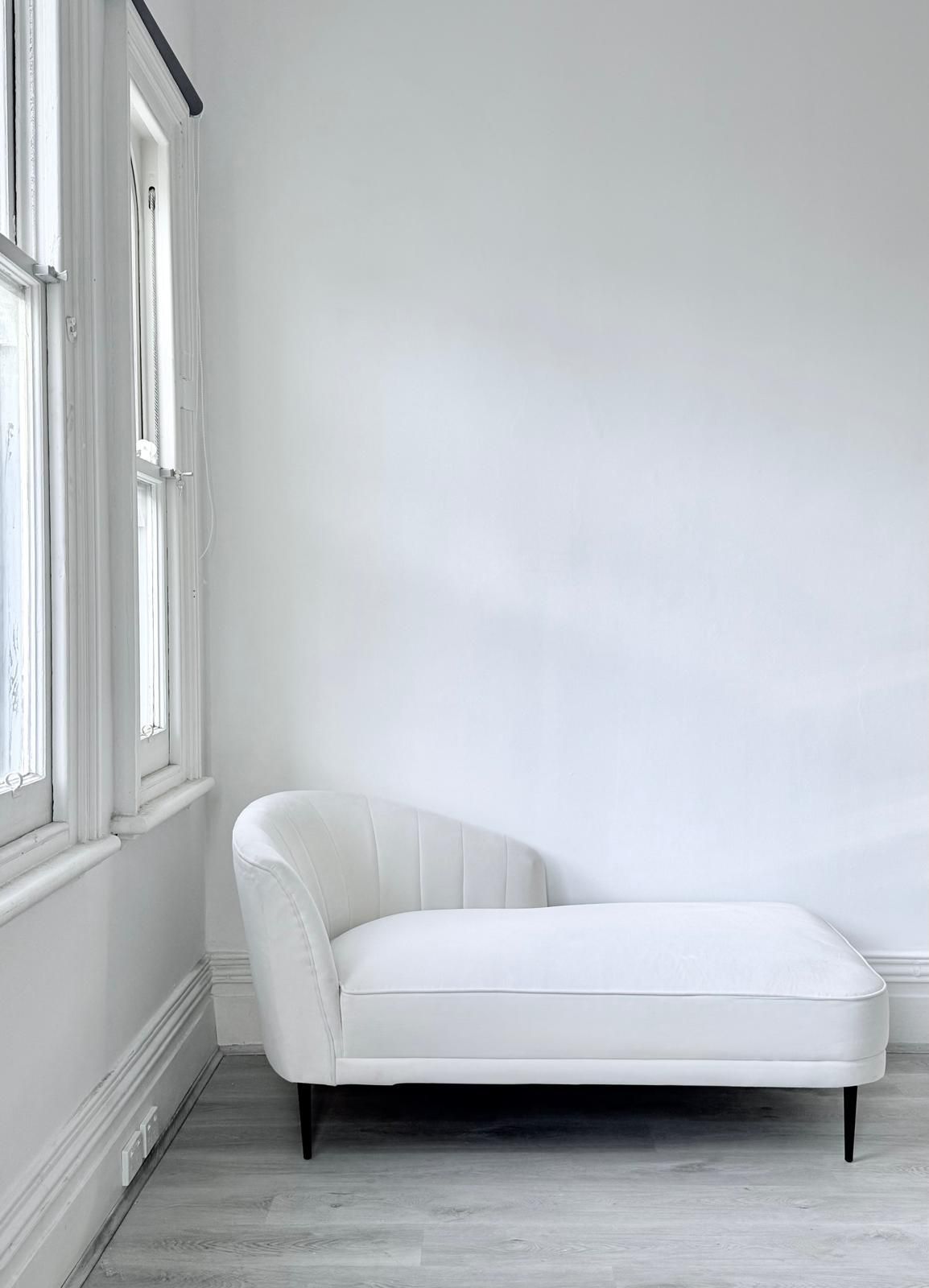 Minimalist white room interior with a modern chaise lounge sofa against white walls and large windows.